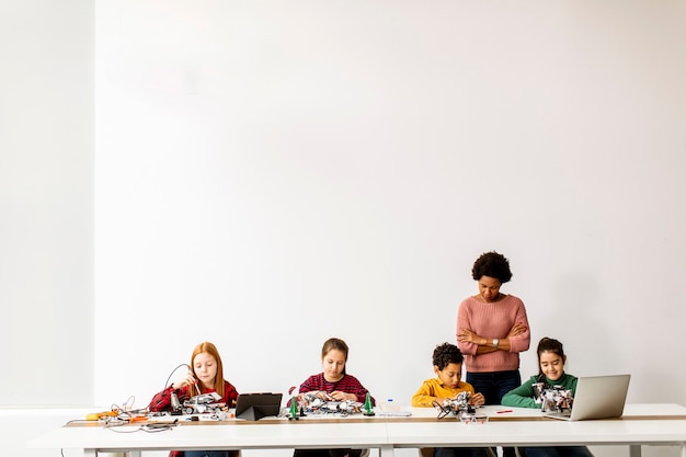 Gruppe glücklicher Kinder mit ihrer afroamerikanischen Wissenschaftslehrerin mit Laptop, der elektrisches Spielzeug und Roboter am Robotikklassenzimmer programmiert