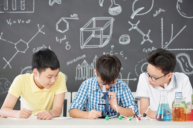 Gruppe intelligenter asiatischer Schulkinder, die im Chemieunterricht mit Mikroskop arbeiten