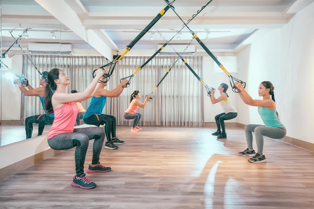 Gruppe junger Asiaten, die Kniebeugen mit Fitness-Aufhängungsgurten machen. gesunde sportliche freunde trainieren in einem sonnigen hellen fitnessstudio. fünf schüler in der trainingsstunde konzentrierten sich auf übungen.