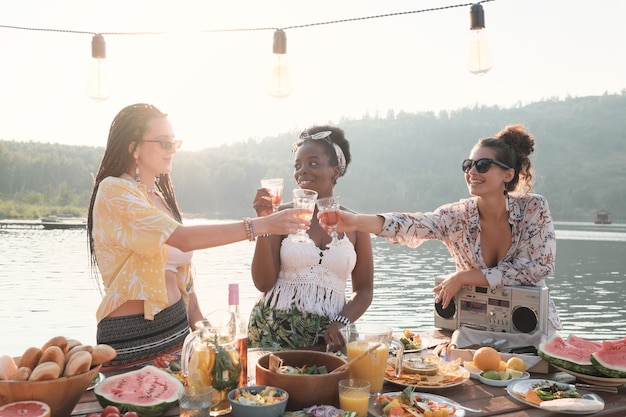 Gruppe junger Frauen, die mit Gläsern Wein rösten, während sie auf der Natur zu Abend essen