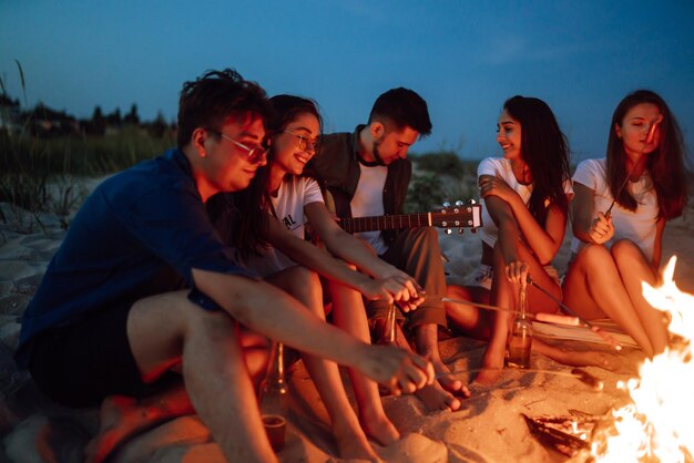 Gruppe junger Freunde, die am Strand sitzen und Würstchen braten Ein Mann spielt Gitarre Campingzeit