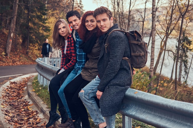 Gruppe junger Freunde mit Rucksäcken, die auf Leitplanken in der Nähe der Straße sitzen, mit einem schönen Wald und Fluss im Hintergrund. Reisen, Wandern, Konzept.