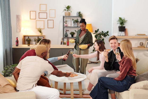 Gruppe junger, fröhlicher interkultureller Freunde in eleganter Freizeitkleidung, die an einem kleinen weißen runden Tisch im Wohnzimmer sitzt, Pizza und Getränke isst