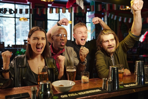 Gruppe junger interkultureller ekstatischer Fußballfans, die ihre Zufriedenheit mit ihrer Lieblingsfußballmannschaft während der Übertragung in einer modernen Kneipe ausdrücken