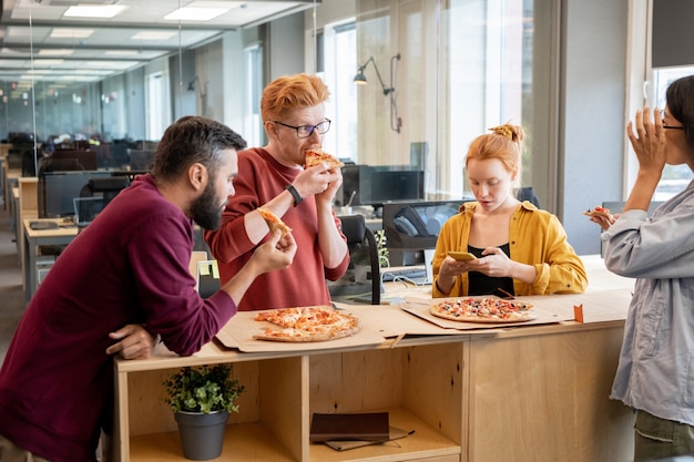 Gruppe junger Kollegen in Freizeitkleidung, die in der Mittagspause mit dem Smartphone reden und sie benutzen, während sie Pizza am Tisch in einem großen zeitgenössischen Büro essen