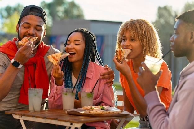 Gruppe junger, lächelnder, stilvoller afroamerikanischer Freunde, die im Café Pizza essen und Limonade trinken