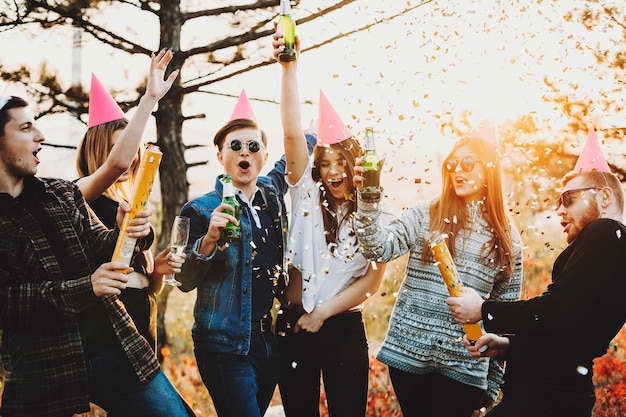 Foto gruppe junger leute, die bierflaschen und explodierende partycracker mit bunten konfetti anheben, während weihnachten in der natur feiern