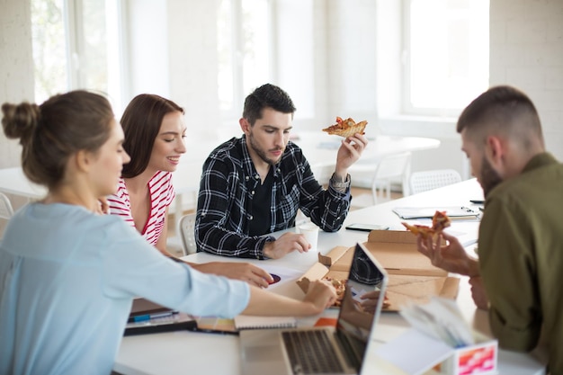 Gruppe junger Männer und Frauen, die in einem modernen Büro glücklich zusammen Pizza essen Junge kreative Menschen verbringen Zeit bei der Arbeit