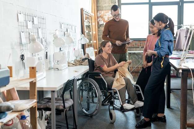 Gruppe junger Modedesigner rund um behinderte Näherin im Rollstuhl während der Diskussion über die Hauptpunkte der neuen Kollektion