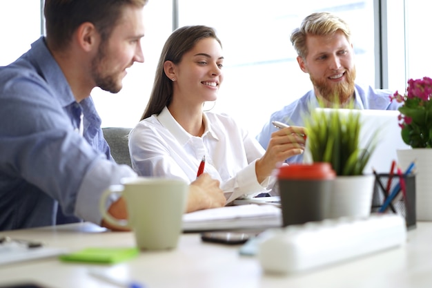 Gruppe junger moderner Menschen in eleganter Freizeitkleidung, die auf den Laptop zeigen und lächeln, während sie im Büro sitzen.