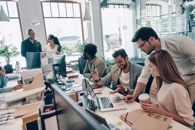 Gruppe junger moderner Menschen in intelligenter Freizeitkleidung, die während der Arbeit im Büro kommunizieren und moderne Technologien verwenden