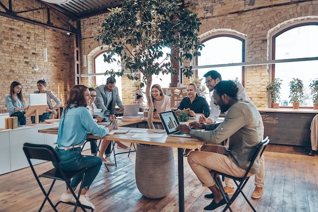 Gruppe junger moderner Menschen in intelligenter Freizeitkleidung, die während der Arbeit im Büro kommunizieren und moderne Technologien verwenden