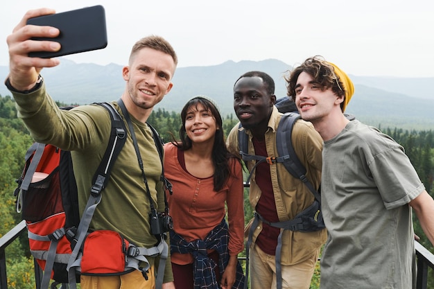 Gruppe lächelnder junger multiethnischer Wanderfreunde, die währenddessen für ein Selfie vor der Waldlandschaft posieren