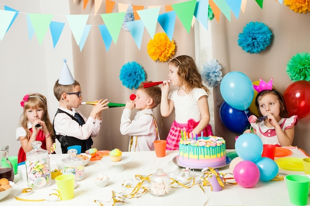 Gruppe lächelnder Kinder, die auf der Geburtstagsfeier im dekorierten Raum spielen. Glückliche Kinder, die auf der Geburtstagsfeier in Pfeifen blasen