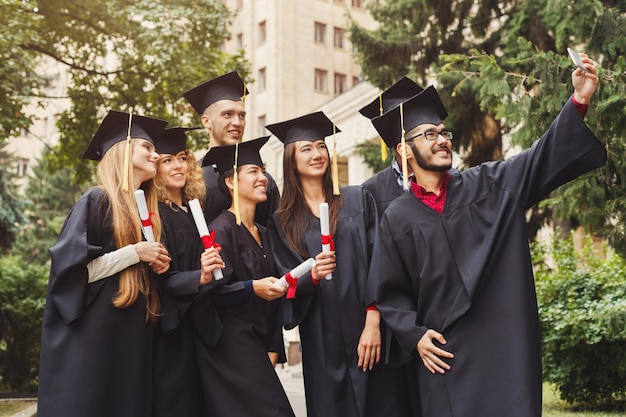 Gruppe multiethnischer Studenten, die sich mit dem Smartphone selbst machen, um ihren Abschluss zu feiern. Bildungs-, Qualifikations- und Kleiderkonzept.