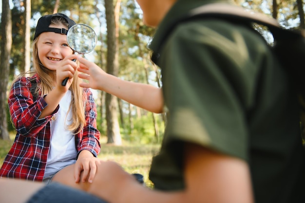 Foto gruppe neugieriger glücklicher schulkinder in freizeitkleidung mit rucksäcken, die gemeinsam natur und wald an einem sonnigen herbsttag erkunden mädchen mit lupe und blick auf tannenzapfen in jungenhänden