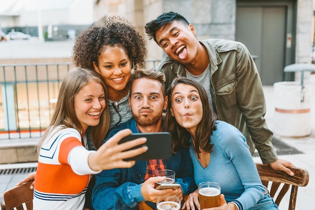 Gruppe positiver gemischtrassiger Freunde, die lustige Gesichter machen, während sie am Café-Tisch ein Selbstporträt auf dem Smartphone machen