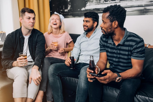 Gruppe verschiedener fröhlicher Freunde, die Spaß bei der Party zu Hause haben, fröhlich lachen, Getränke genießen, die auf dem Sofa im Wohnzimmer sitzen. Konzept für Menschen, Freizeit und Freundschaft.