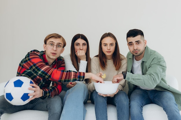 Gruppe von 4 Freunden, die sich zu Hause entspannen, Chips essen und wichtige Fußballspiele im Fernsehen ansehen Junge Freunde sitzen mit Chips auf dem Sofa und beobachten angespannte Momente im Fußballspiel