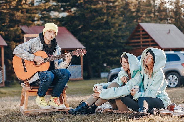 Gruppe von Freunden beim musikalischen Abend im Innenhof des Camps bei Sonnenuntergang die Atmosphäre genießen. Der Typ auf dem Hocker spielt eine Akustikgitarre. Die Mädchen sitzen nebeneinander und hören zu.