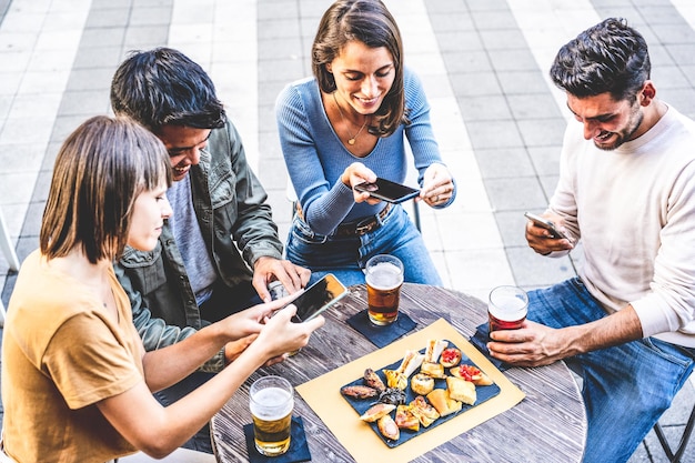 Gruppe von Freunden der Generation z in Kneipen, die Spaß haben und Smartphones verwenden, um glückliche Studenten mit Speisen und Getränken zu fotografieren, die eine App verwenden, um Inhalte in sozialen Medien zu teilen