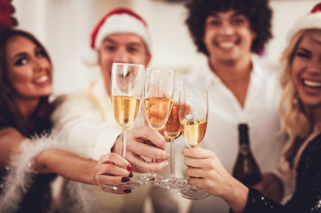 Gruppe von Freunden, die zu Hause Weihnachten feiern. Sie haben Spaß und stoßen mit Champagner an. Selektiver Fokus. Konzentrieren Sie sich auf den Vordergrund, auf Gläser Weißwein.