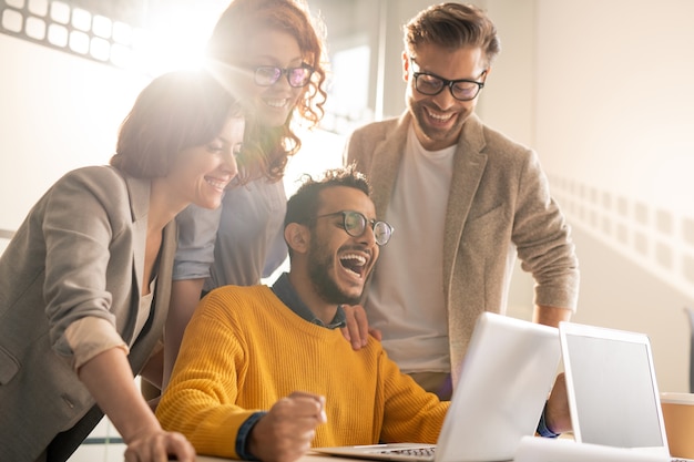 Gruppe von fröhlichen aufgeregten jungen interracial Kollegen, die lachen, während sie Internetnachrichten auf Laptop lesen