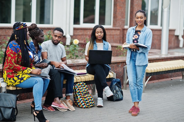 Gruppe von fünf afrikanischen College-Studenten, die gemeinsam Zeit auf dem Campus des Universitätshofs verbringen Schwarze Afro-Freunde, die an der Bank mit Schulartikeln, Laptops, Notizbüchern studieren