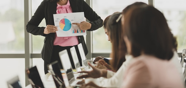 Gruppe von Geschäftsfrauen, die sich im Büro treffen, Führer oder Manager, die Diagramm und Diagramm halten, während sie dem Publikum Details und Bedeutung erklären, die Kollegen hören ihr zu und drücken ihr Verständnis aus.