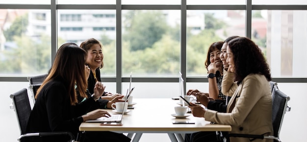 Gruppe von Geschäftsfrauen in formeller Geschäftskleidung, die zusammen am Besprechungstisch sitzen und auf Laptop-Computer und Touchscreen-Tablet tippen, unterhalten sich im Konferenzraum in der Nähe von Glasfenstern.