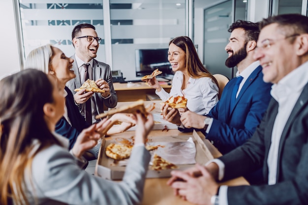 Gruppe von Geschäftsleuten, die im Sitzungssaal sitzen, plaudern, lachen und Pizza zum Mittagessen haben.