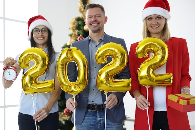 Gruppe von Geschäftsleuten in Weihnachtsmann-Hüten, die goldene Ballons mit den Nummern 2022 in der Nähe des Baumes des neuen Jahres halten. Firmenkonzept für Neujahrsferien