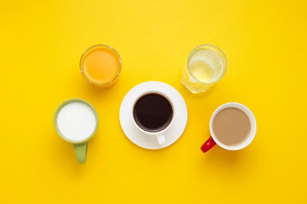 Gruppe von Getränken in bunten Tassen, schwarzer Kaffee, Kaffee mit Milch, Joghurt, nur Wasser, Orangensaft auf gelbem Hintergrund. Flache Lage, Draufsicht