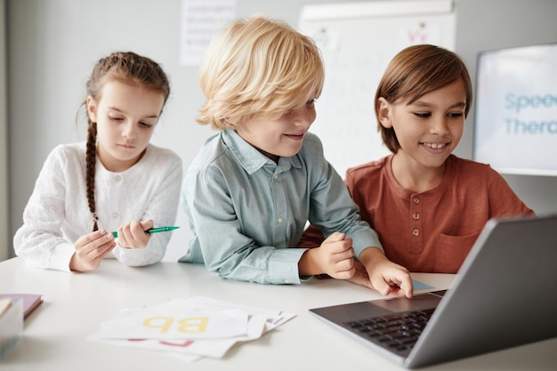 Gruppe von Kindern, die während einer Lektion, die sie online lernen, am Tisch sitzen und auf dem Laptop tippen