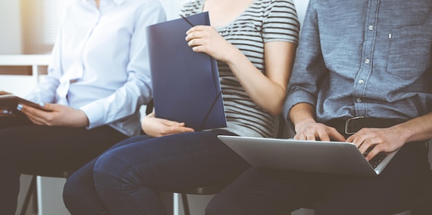 Gruppe von lässig gekleideten Geschäftsleuten, die in einer Schlange sitzen und auf ein Vorstellungsgespräch im sonnigen Büro warten