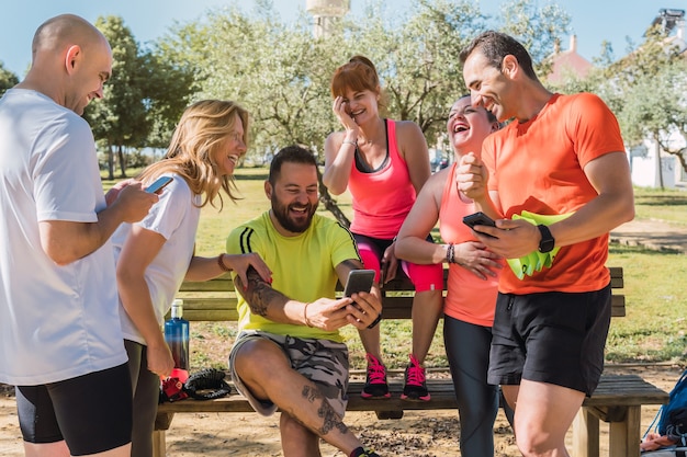 Gruppe von Leuten in Sportkleidung, die lachen, während sie das Handy in einem Park benutzen