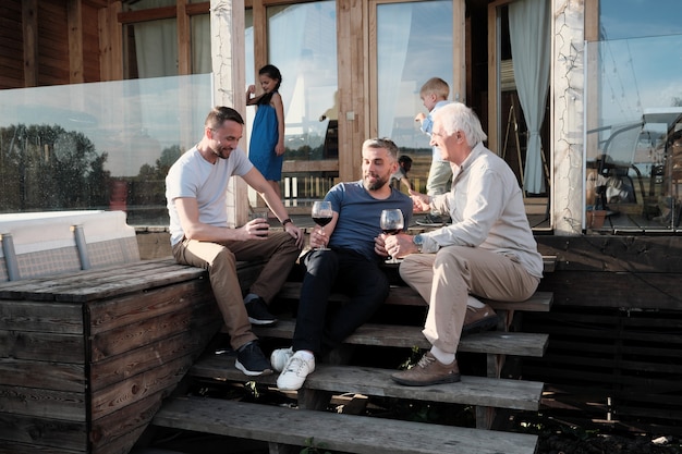 Gruppe von Männern, die auf der Veranda sitzen, miteinander reden und Rotwein trinken, den sie auf dem Land draußen ruhen