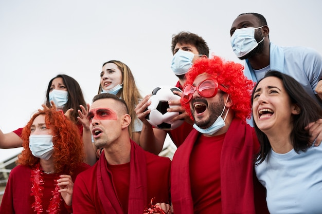 Gruppe von Menschen verschiedener Rassen, die ihr Team im Fußballstadion mit Gesichtsmasken aufmuntern