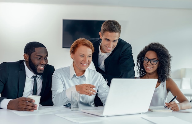 Gruppe von Mitarbeitern in Business-Freizeitkleidung, die zusammen am Tisch sitzen und auf den Bildschirm des Laptops schauen