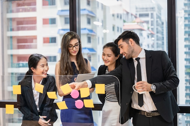 Gruppe von multiethnischen Geschäftskollegen, die Geschäftsideen mit Haftnotiz an Bord im modernen Büro analysieren und diskutieren