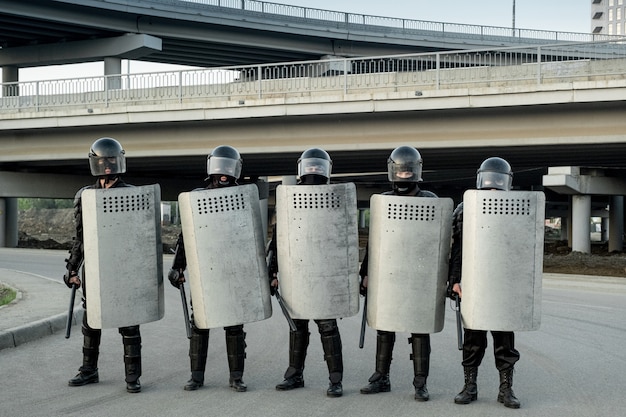 Gruppe von Polizeiwachen in Helmen, die Schlagstöcke und Schutzschilde mit seitlichem Griff halten und unter Brücken in Reihe stehen