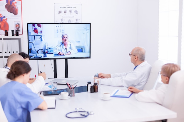 Gruppe von Ärzten, die während des Videoanrufs im Konferenzraum medizinischen Experten zuhören. Medizinpersonal, das das Internet während des Online-Meetings mit einem erfahrenen Arzt nutzt, um Fachwissen zu erhalten.