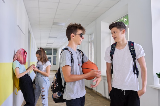 Gruppe von Teenagern, die sich unterhalten, stehen vor dem Schulgebäude, zwei Männer mit Rucksäcken, Basketballball im Fokus, Schulmädchen im Korridorhintergrund. Lifestyle, Kommunikation, Jugendkonzept