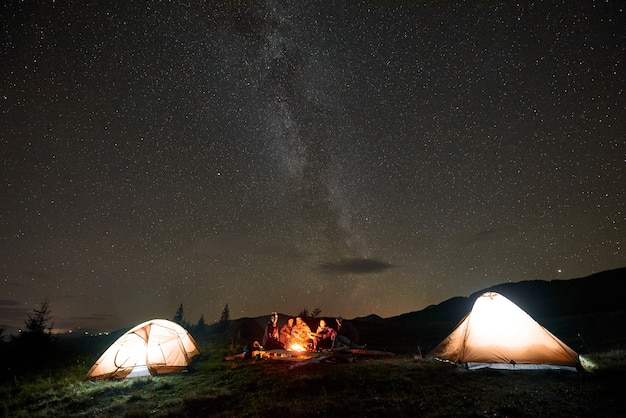 Gruppe von Touristen mit Gitarre durch brennendes Lagerfeuer unter dunklem Sternenhimmel mit Milchstraßenkonstellation.