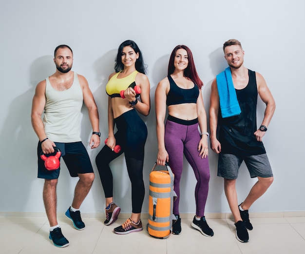 Gruppe von vier sportlichen jungen Freunden, die zusammen im Fitnessstudio an der grauen Wand stehen