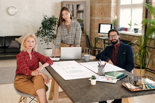 Gruppe von zwei eleganten Frauen und jungen bärtigen Geschäftsmann, die Sie beim Arbeitstreffen am großen Holztisch im Büro ansehen