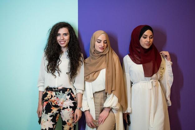 Gruppenporträt schöner muslimischer Frauen, zwei davon in modischer Kleidung mit Hijab, isoliert auf buntem Hintergrund, die moderne islamische Mode und das Ramadan-Kareem-Konzept repräsentieren.