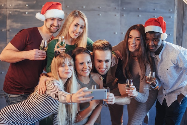 Gruppieren Sie die schönen jungen Leute, die selfie in der Partei des neuen Jahres, in den Mädchen und in den Jungen der besten Freunde tun, die zusammen Spaß haben und emotionale Lebensstilleute aufwerfen. Hüte Santas und Champagnergläser in ihren Händen
