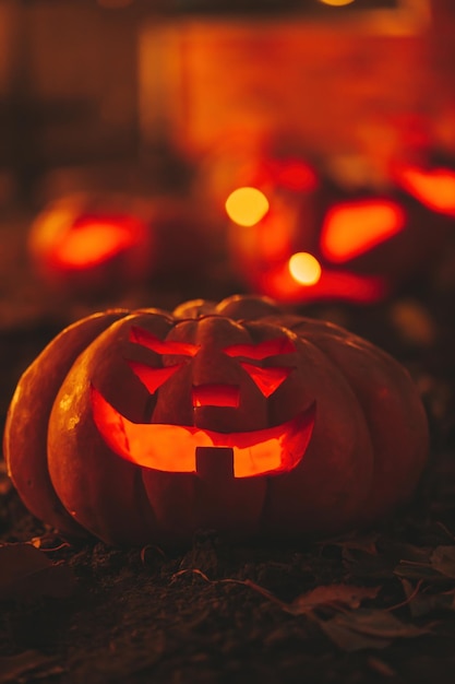 Gruselige Jack-Laterne-Halloween-Kürbisse in der Dunkelheit auf dem Boden zwischen trockenen Blättern auf der Straße. Dekoration am Heiligabend. Lustiger leuchtender Kürbis mit Kerzen auf dunklem Hintergrund im Freien in der Nähe des Hauses