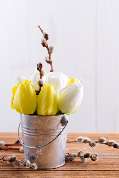 Grußkarte mit frischen Frühlingstulpenblumen und frischen Weidenzweigen in einem Eimer auf einem braunen Holztisch.